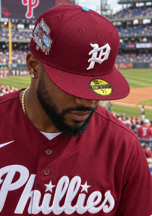 A man wearing a Philadelphia Phillies baseball cap and matching maroon jersey stands in a stadium. The cap features a white 'P' logo and a commemorative patch, while the jersey displays the team name 'Phillies' in white script.