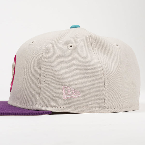 A structured baseball cap in light gray with a purple flat brim, featuring a pink embroidered New Era flag logo on the side and a small teal button on top.